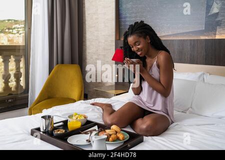Tout le contenu jeune afro-américaine touristique dans vêtements de nuit sur un lit confortable avec plateau de délicieux petit déjeuner et boire du café Banque D'Images