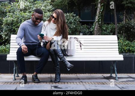 Ensemble complet de couple multiethnique heureux avec des tenues élégantes assis sur banc et en parcourant votre téléphone mobile ensemble tout en passant du temps dans le parc Banque D'Images