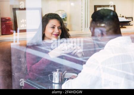 Bonne femme avec une tasse de boisson chaude appréciant une conversation agréable avec un petit ami noir tout en prenant le petit déjeuner ensemble dans un café confortable Banque D'Images
