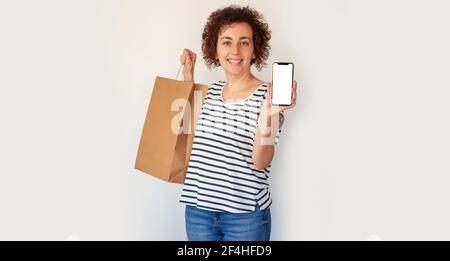 Une femme caucasienne aux cheveux maurires montre son téléphone portable à l'appareil photo tout en tenant les sacs d'achats sur un fond blanc avec espace de copie Banque D'Images