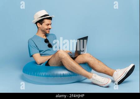 Joyeux souriant caucasien freelance ou étudiant portant un chapeau assis sur anneau gonflable de natation, en utilisant un ordinateur portable pour le travail en ligne ou l'apprentissage à distance pendant les vacances, isolé fond bleu Banque D'Images