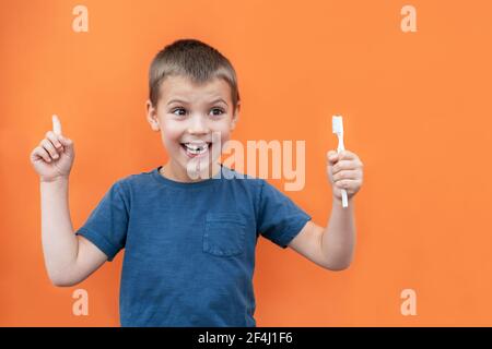 Un garçon sans la dent supérieure de lait dans un t-shirt bleu tient la brosse à dents à la main sur fond orange. Banque D'Images