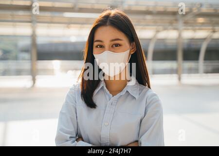 Portrait d'une jeune femme d'affaires asiatique portant un masque de protection Covid-19 pour la sécurité contre le coronavirus avec les armes croisées à l'extérieur du bureau Banque D'Images