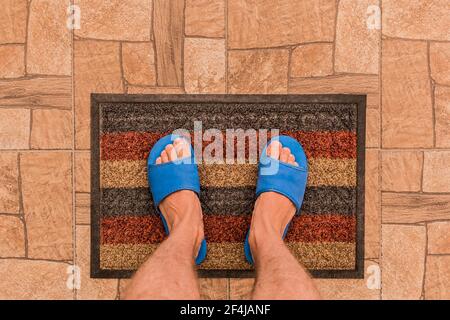 Les pieds mâles dans des pantoufles maison bleues se tiennent sur un tapis de pied sur un fond de sol carrelé brun texture, vue de dessus. Banque D'Images