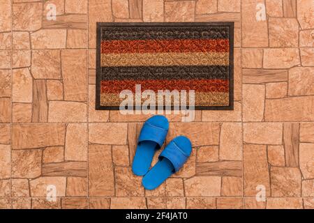 Les pantoufles maison bleues pour hommes se tiennent près du tapis de pied sur un fond de sol carrelé marron texturé, vue du dessus. Banque D'Images