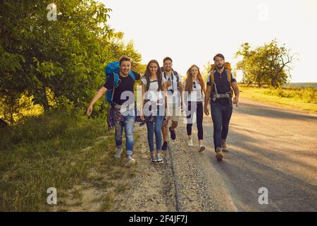 Groupe de jeunes amis heureux avec des sacs à dos marchant le long de la route dans la campagne. Randonneurs en voyage touristique en été Banque D'Images