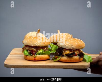 Délicieux hamburger maison frais. Main tenant et montrant quatre délicieux hamburgers faits maison sur plateau en bois ou planche à découper en bois sur fond sombre. Banque D'Images