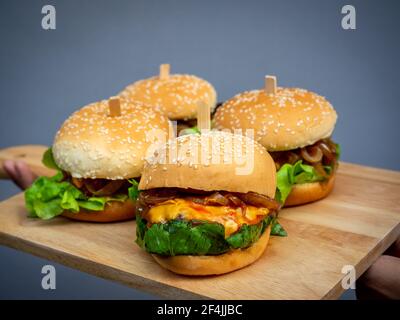 Délicieux hamburger maison frais. Main tenant et montrant quatre délicieux hamburgers faits maison sur plateau en bois ou planche à découper en bois sur fond sombre. Banque D'Images