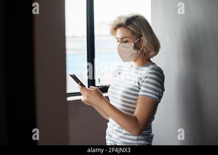 Une dame blonde avec un masque médical discute de la pose blonde près de la fenêtre dans un t-shirt Banque D'Images