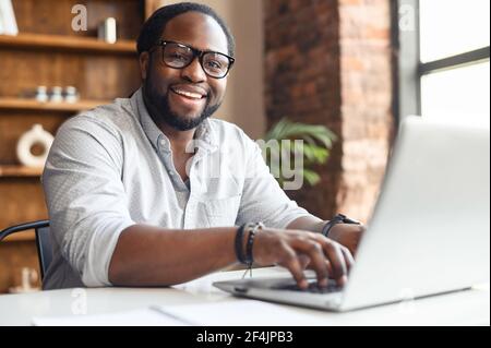 Un blogueur d'employés afro-américains heureux et positif ou UN INFORMATICIEN en lunettes assis à la table avec un ordinateur portable et un dactylographie, écrivant un article, souriant et regardant un appareil photo, aime travailler dans son entreprise Banque D'Images