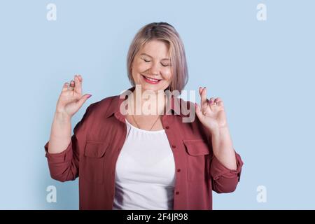 La femme âgée de race blanche aux cheveux blonds pense à quelque chose sourire et croiser les doigts sur un mur bleu de studio Banque D'Images