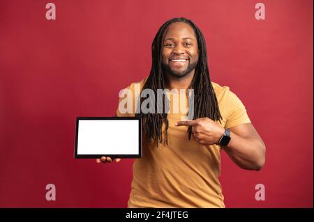 Homme noir de race mixte avec des dreadlocks se tenant isolé sur fond rouge, présentant une nouvelle application, pointant avec le doigt sur la tablette numérique avec espace blanc pour la publicité, image maquette Banque D'Images