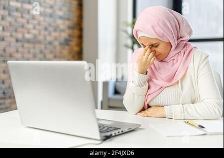 Une femme d'affaires arabe de race musulmane épuisée ou une étudiante dans le hijab a fermé les yeux touchant le front souffrant de maux de tête graves migraines, se sentant fatiguée submergée par le travail informatique Banque D'Images