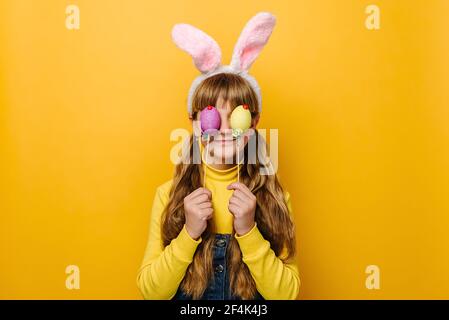 Portrait de petite fille drôle tient et couvre les yeux avec deux oeufs de couleur de Pâques, porte des oreilles roses de lapin moelleux, isolé sur fond jaune studio Banque D'Images