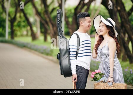 Beau jeune homme avec guitare regardant heureux rire joli petite amie quand ils sont debout dans le parc Banque D'Images