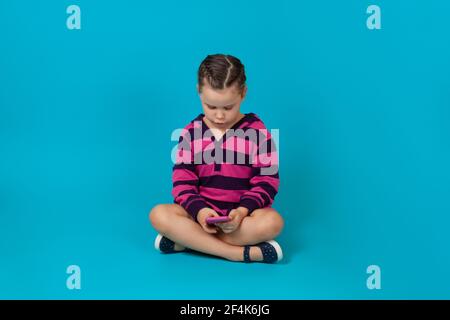 portrait d'une jeune fille enthousiaste à capuche rayée, assise à pattes croisées sur le sol et jouant à des jeux sur un smartphone, isolée sur un fond bleu Banque D'Images