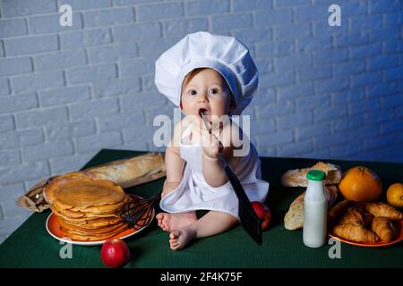 Petit beau enfant habillé dans un tablier, costume de cuisine pour enfants. Crêpes et pâtisseries fraîches sur la table Banque D'Images