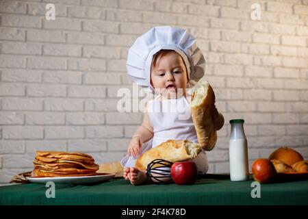 Petit garçon habillé comme un cuisinier dans la cuisine. Beau enfant habillé dans un tablier mangeant du pain Banque D'Images