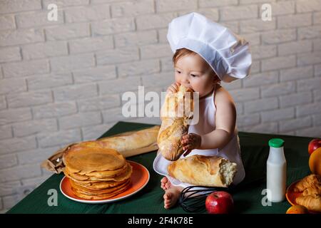 Petit garçon habillé comme un cuisinier dans la cuisine. Beau enfant habillé dans un tablier mangeant du pain Banque D'Images