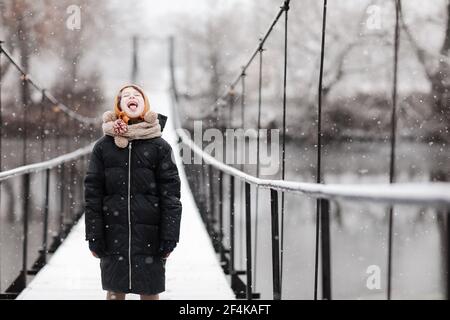 Une petite fille drôle attrape des flocons de neige dans un beau parc d'hiver pendant une chute de neige. Bébé mignon joue dans la neige. Activités d'hiver pour les enfants Banque D'Images