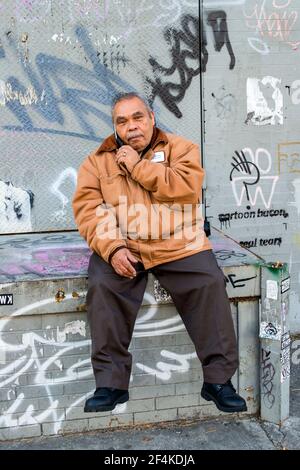 New York City, USA. Hispanic man making a molbile appel alors qu'il était assis en face d'un mur infestés graffity. Banque D'Images