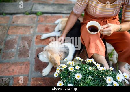 Femme âgée méconnue avec café assis à l'extérieur dans le jardin, amitié avec un chien d'animal de compagnie. Banque D'Images