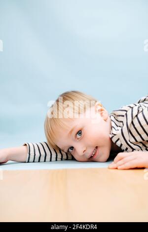 Beau garçon blond allongé sur le sol, jouant mignon. Il porte une chemise rayée à manches longues. Prise de vue en studio. Sur fond bleu. Regarder la caméra. Banque D'Images