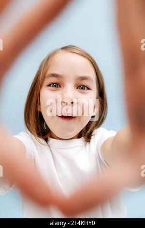 Drôle souriant fille de 9 ans à travers un cadre de doigt elle fait. Sur fond bleu. Prise de vue en studio. Elle ouvre sa bouche, les dents couvertes de plié Banque D'Images