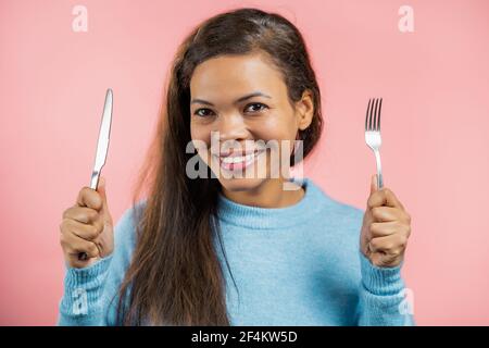 Portrait d'une femme affamée avec fourchette et couteau. Femme d'âge mûr attendant de servir des plats de dîner avec des couverts sur fond rose studio. Banque D'Images