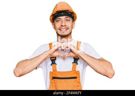 Un jeune homme hispanique portant un uniforme de handyman et un casque de sécurité souriant dans l'amour montrant le symbole du cœur et la forme avec les mains. Concept romantique. Banque D'Images