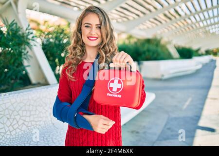 Jeune fille blonde souriante heureuse blessée avec harnais de bras et tenue de trousse de premiers soins au parc. Banque D'Images