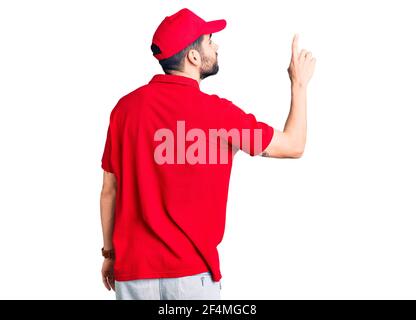 Jeune homme beau avec barbe portant l'uniforme de livraison posant en arrière en pointant vers l'avant avec le doigt Banque D'Images