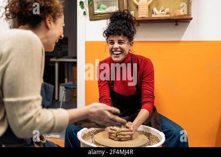 Photo de stock de femmes heureuses en tablier travaillant derrière une roue de potier dans un atelier. Banque D'Images