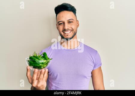 Jeune homme arabe tenant un bol de poivrons verts ayant l'air positif et se tenir debout et sourire avec un sourire confiant dents Banque D'Images