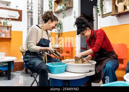 Photo de stock de femmes heureuses en tablier travaillant derrière une roue de potier dans un atelier. Banque D'Images