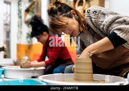 Photo de la femme concentrée dans un tablier travaillant derrière une roue de potier dans un atelier. Banque D'Images