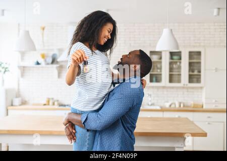 Un jeune couple afro-américain hilarant célébrant l'achat d'une propriété, un homme excité portant une femme joyeuse avec des clés dans sa main, les jeunes mariés tiennent un porte-clés sous la forme d'une maison mignonne Banque D'Images