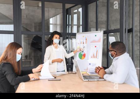 Équipe de travail diversifiée dans les masques médicaux de protection travaillant ensemble dans l'espace de travail de collègue moderne, remue-méninges multiraciaux employés pendant la période pandémique, une femme biraciale présentant des idées. Nouvelles règles sociales Banque D'Images