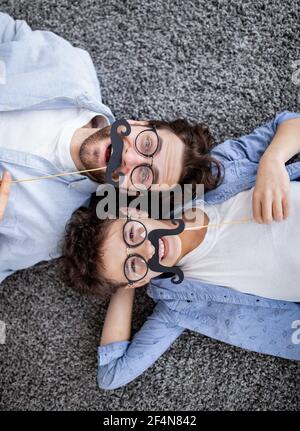 Joyeux père et son fils s'amusant avec une fausse moustache sur des bâtons, portant des lunettes et souriant à l'appareil photo, vue du dessus Banque D'Images