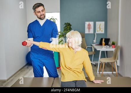 Une femme âgée lève une haltère, faisant un exercice de traitement avec son physiothérapeute. Traitement physiologique dans un centre de réadaptation Banque D'Images