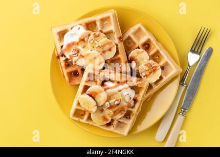 Gaufres belges avec banane, sauce caramel et amandes sur plaque jaune, fond jaune. Vue de dessus. Petit déjeuner sucré avec gaufres à la banane Banque D'Images