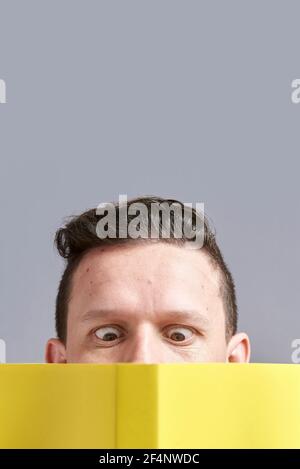 Jeune homme lisant un livre jaune avec les yeux accroués; un regard comique et drôle. Portrait frontal avec arrière-plan gris et espace de copie. Banque D'Images