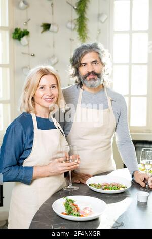 Homme et femme matures en tabliers debout près d'une table de cuisine servi avec des plats maison appétissants sur des assiettes et du vin blanc dans les goujures Banque D'Images