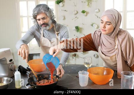 Jeune femme musulmane versant du ketchup de tomate dans de la viande hachée poêle à frire tout en aidant l'homme barbu à cuire le remplissage pour la pâtisserie Banque D'Images