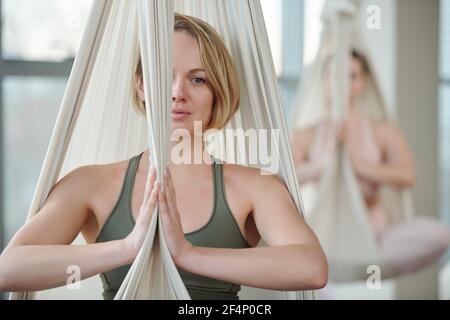 Jeune femme en survêtement gris, assise en posture de lotus avec ses mains ensemble par la poitrine tout en faisant de l'exercice en salle de sport contre son ami Banque D'Images