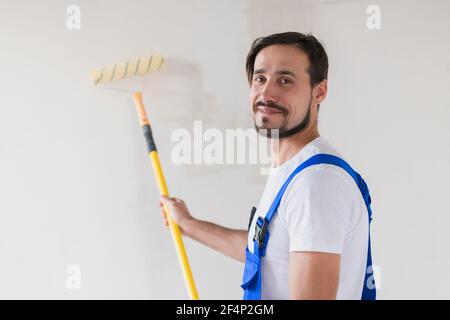 Réparateur peint le mur avec un rouleau, sourit et regarde la caméra Banque D'Images