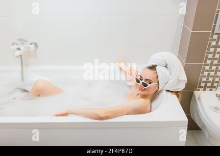 Jolie femme avec un sourire blanc neige dans la salle de bains. Femme élégante dans une serviette sur la tête et lunettes de soleil prend des soins spa à la maison ou à l'hôtel Banque D'Images