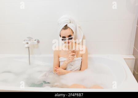 Jolie femme avec un sourire blanc neige dans la salle de bains. Femme élégante dans une serviette sur la tête et lunettes de soleil prend des soins spa à la maison ou à l'hôtel Banque D'Images