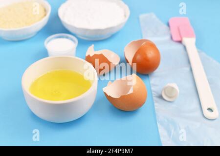 Ingrédients pour faire des macarons français faits maison avec un bol à œuf blanc à côté de coquilles d'œufs cassées sur fond bleu Banque D'Images