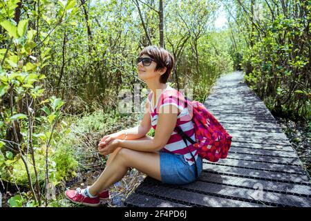 Une femme avec sac à dos assise sur le chemin en bois et se détendre après la randonnée dans la forêt d'été. Apprécier, unité avec la nature, détox numérique Banque D'Images